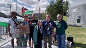 6 people, some with scarves and signs, standing in front of Palestinian flags.