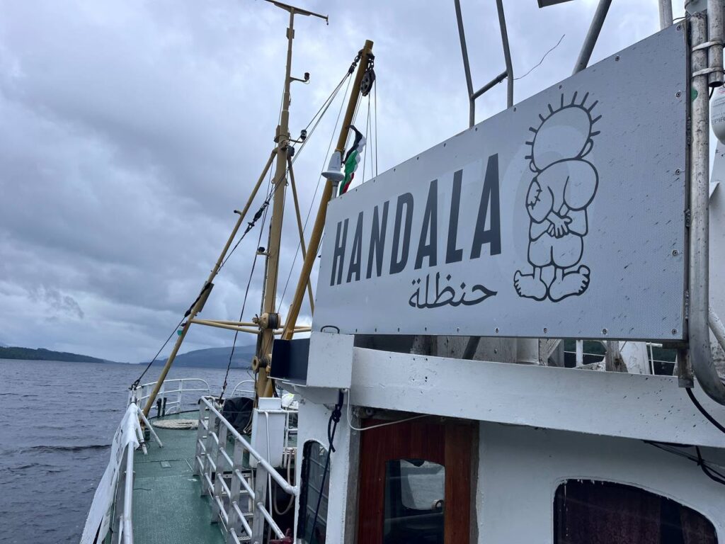 Partial shot of deck and rail of the boat, with large banner of "Handala" (in English and Arabic) with the cartoon figure of Handala.