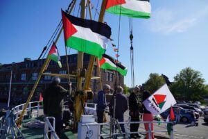 People standing on bow of 'Handala' boat in port, with Palestinian flags, and a flag reading "Ship to Gaza".